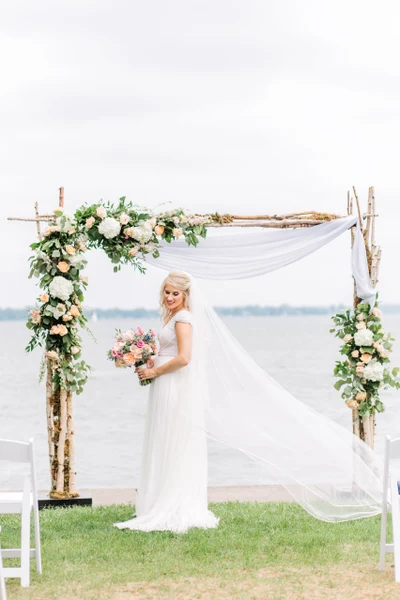 Rose's Bouquets flower and birch ceremony arch with white draping beside a lake
