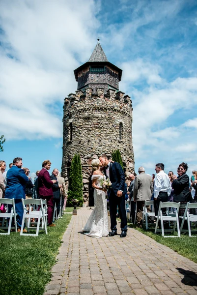 Crown Coordination couple kissing at an outdoor ceremony in front of a stone tower with guests seated on the lawn