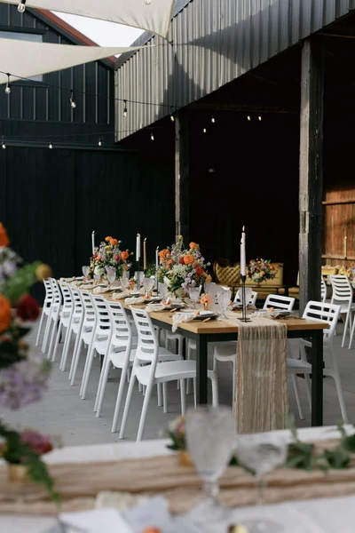 RD Events & Design long dinner tables with white chairs and bright flowers in a barn courtyard under string lights
