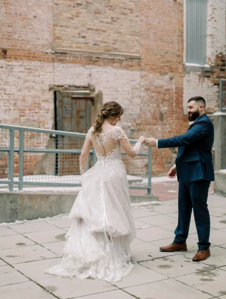 J.Luxe Salon bride with a half-up curled style holding hands with the groom beside a brick building