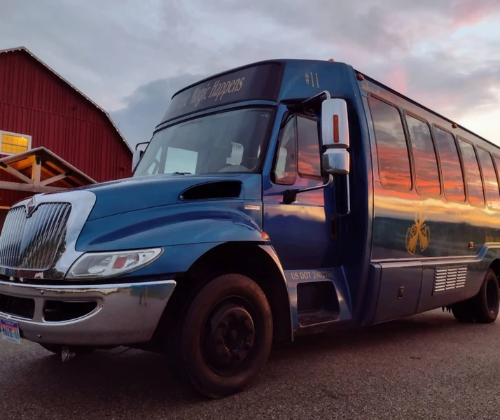 The Magic Shuttle Bus blue coach parked beside a red barn at sunset