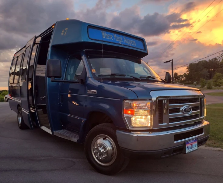 The Magic Shuttle Bus blue shuttle bus with its door open under a sunset sky