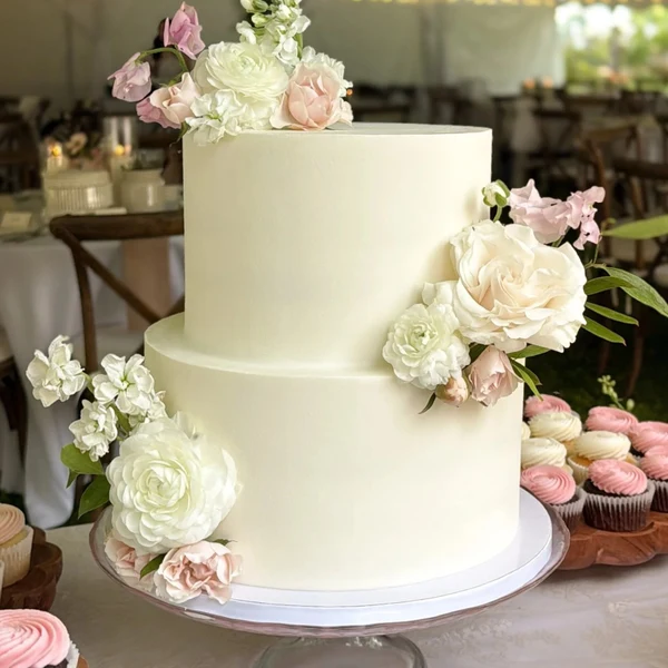 Butter & Batter Bakery two-tier white wedding cake with fresh flowers beside cupcakes under a reception tent