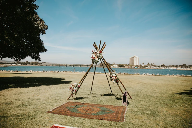 Wonder Event Rentals wooden tipi ceremony arch with flowers and a vintage rug on a bayfront lawn