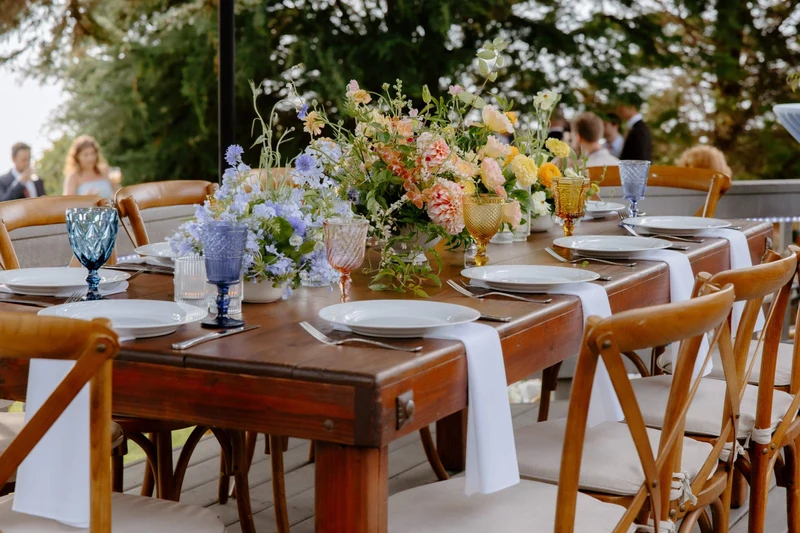 Personal Touch Dining wood farm table set with blue goblets and garden flowers at an outdoor reception