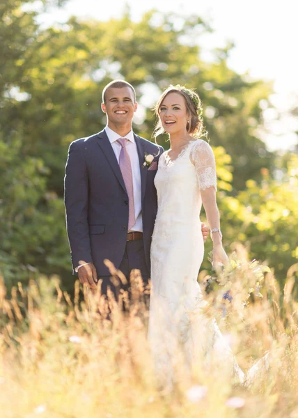 On-Site Bridal bride with soft curls and flowers in her hair beside the groom in a sunlit field